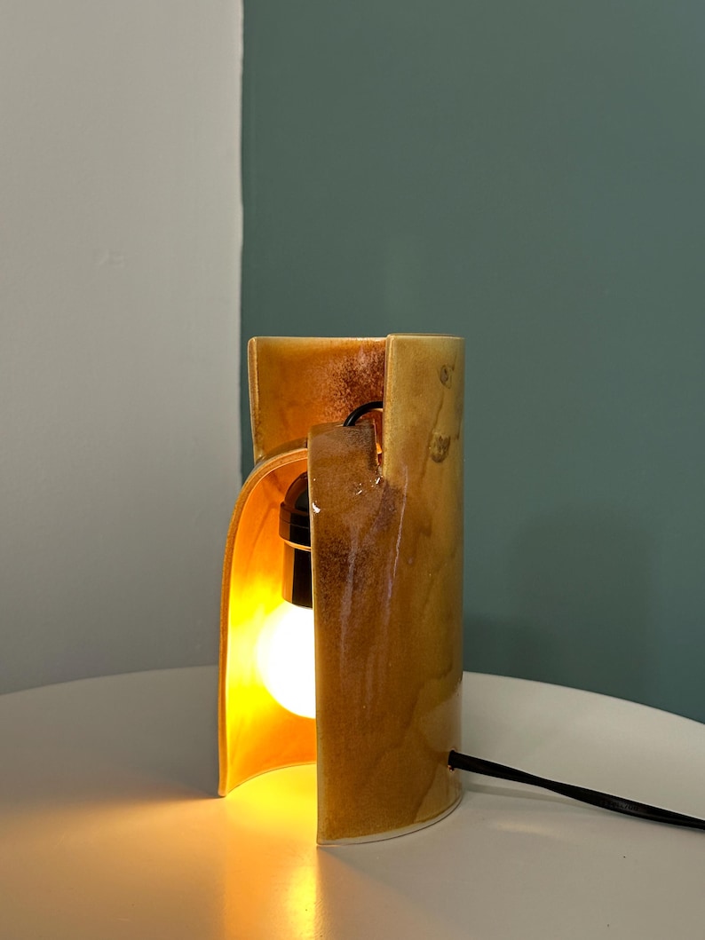 Puede incluir: Una l&aacute;mpara de mesa cil&iacute;ndrica de cer&aacute;mica con un esmalte brillante de tonos &aacute;mbar y marr&oacute;n dorado. La l&aacute;mpara presenta una abertura vertical que revela una bombilla encendida en su interior. Un cable de alimentaci&oacute;n negro sale del lateral.
