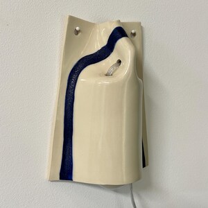 May include: A wall-mounted ceramic light fixture with a glossy cream-colored finish and a single vertical stripe of textured dark blue glaze. The fixture features two circular mounting holes at the top and a central opening for a light bulb.