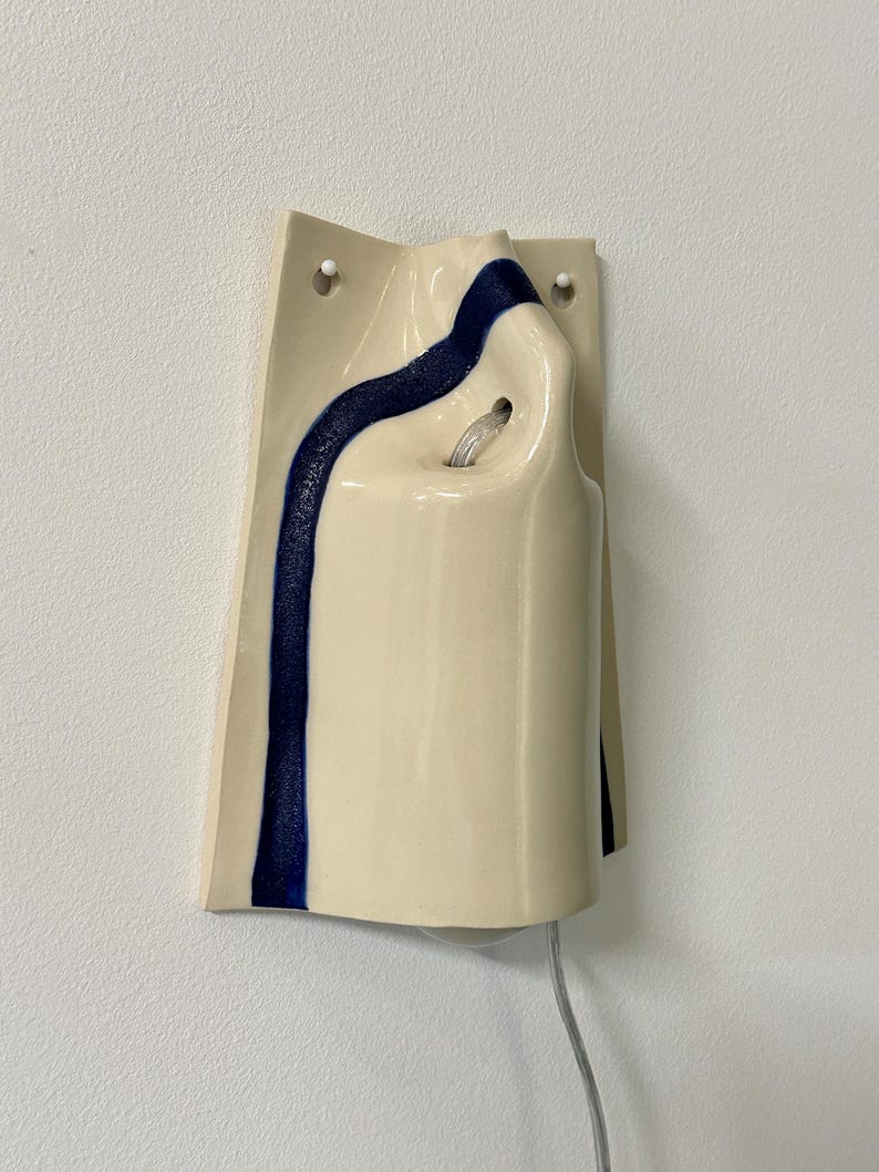 May include: A wall-mounted ceramic light fixture with a glossy cream-colored finish and a single vertical stripe of textured dark blue glaze. The fixture features two circular mounting holes at the top and a central opening for a light bulb.
