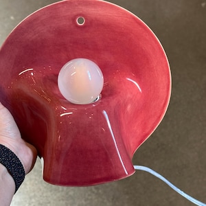 May include: A wall-mounted lamp featuring a glossy, deep red ceramic shade with a wide, flared, organic shape. A single, round, frosted white light bulb is centred within the shade. A small circular hole is visible at the top of the ceramic.