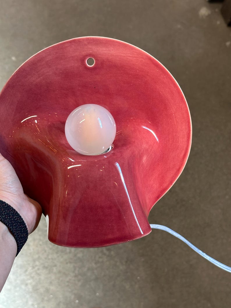 May include: A wall-mounted lamp featuring a glossy, deep red ceramic shade with a wide, flared, organic shape. A single, round, frosted white light bulb is centred within the shade. A small circular hole is visible at the top of the ceramic.