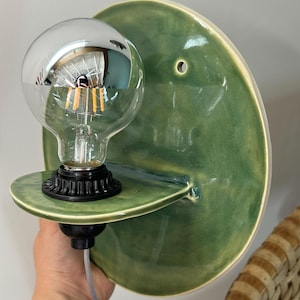 May include: A wall-mounted light fixture featuring a circular, glossy green ceramic backplate with a small mounting hole. A small, semi-circular ceramic shelf extends from the center, holding a black socket with a clear, round LED filament bulb.