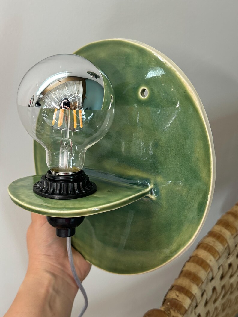 May include: A wall-mounted light fixture featuring a circular, glossy green ceramic backplate with a small mounting hole. A small, semi-circular ceramic shelf extends from the center, holding a black socket with a clear, round LED filament bulb.