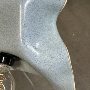 May include: A ceramic lamp shade with a wavy, organic edge and a glossy, speckled light blue finish. The shade features a smooth, curved surface with a subtle gradient of blue tones and a visible light bulb socket attached to the side.