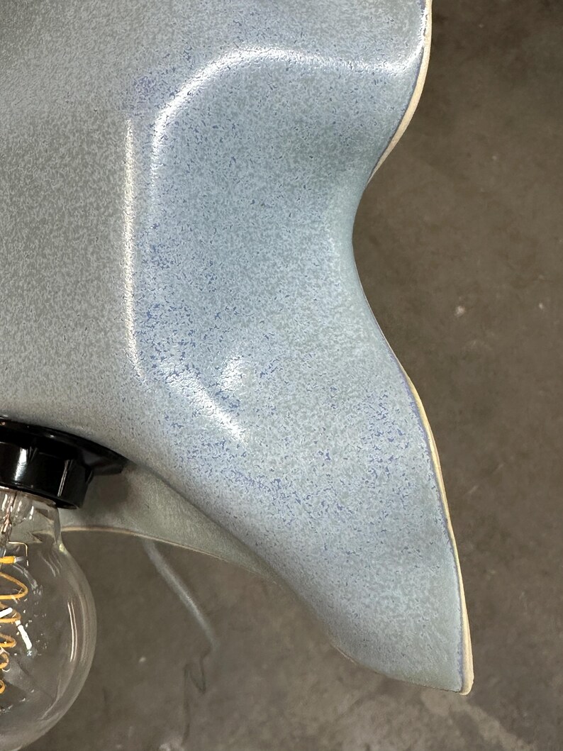 May include: A ceramic lamp shade with a wavy, organic edge and a glossy, speckled light blue finish. The shade features a smooth, curved surface with a subtle gradient of blue tones and a visible light bulb socket attached to the side.