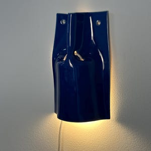 Puede incluir: Un aplique de pared de cer&aacute;mica azul oscuro brillante con una silueta curva y plegada. La l&aacute;mpara cuenta con dos orificios de montaje circulares en la parte superior y emite un brillo c&aacute;lido y suave desde atr&aacute;s, proyectando luz hacia abajo.