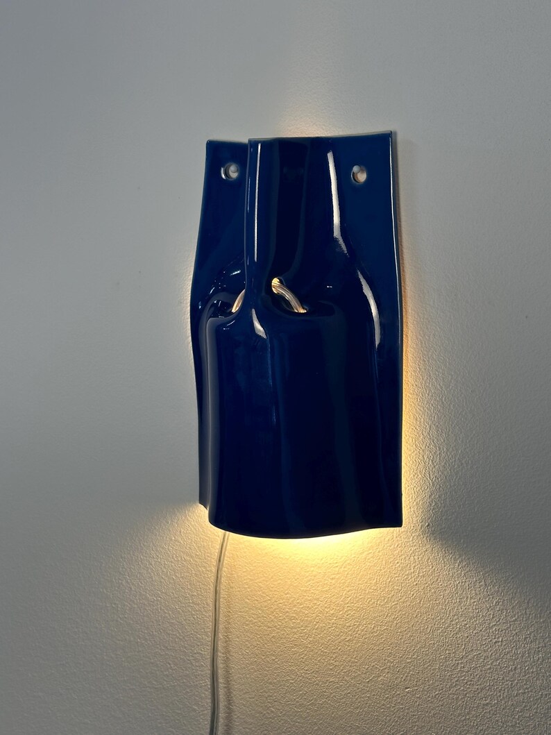 Puede incluir: Un aplique de pared de cer&aacute;mica azul oscuro brillante con una silueta curva y plegada. La l&aacute;mpara cuenta con dos orificios de montaje circulares en la parte superior y emite un brillo c&aacute;lido y suave desde atr&aacute;s, proyectando luz hacia abajo.