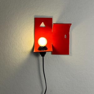 May include: A contemporary wall sconce with a vibrant red geometric design. It features a small, round light bulb and a black cord. The design incorporates a triangle cutout and a rectangular shape.