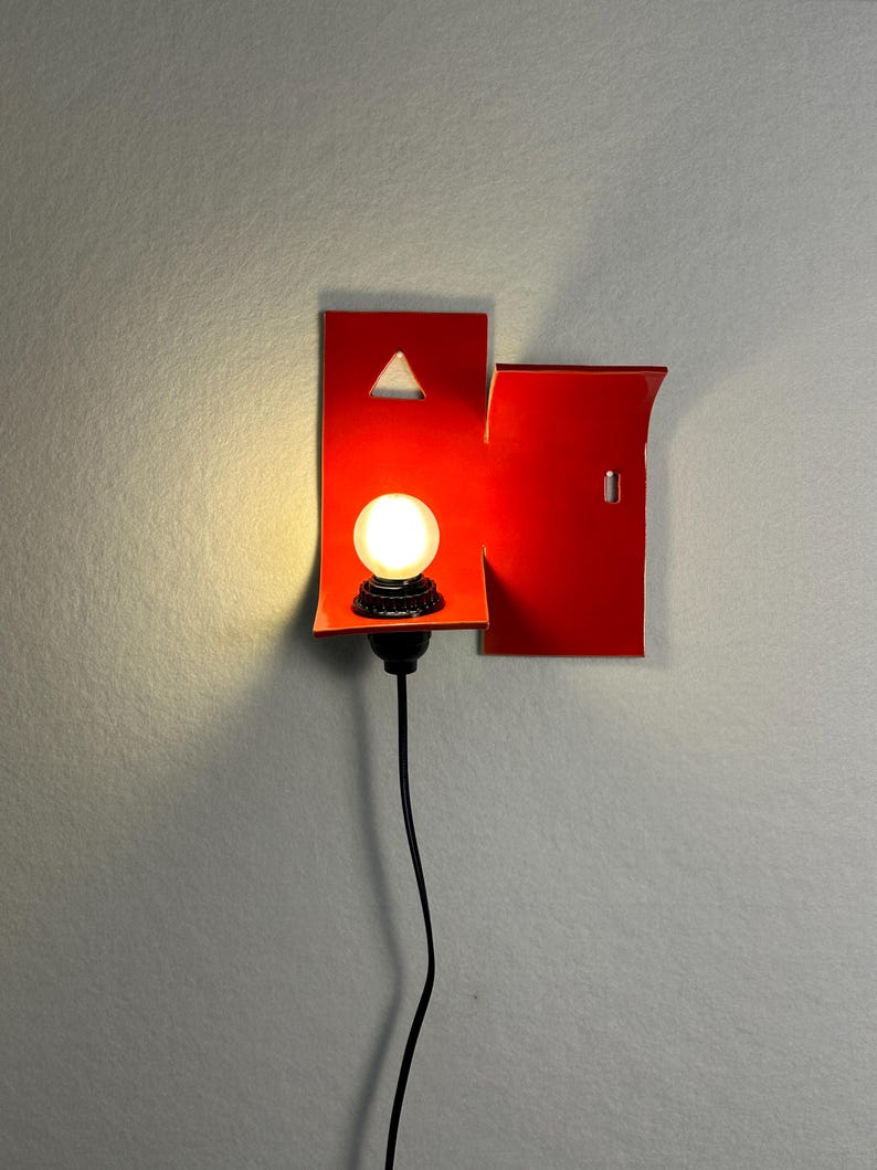 May include: A contemporary wall sconce with a vibrant red geometric design. It features a small, round light bulb and a black cord. The design incorporates a triangle cutout and a rectangular shape.