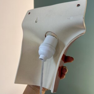Pode incluir: Um candeeiro de parede em cer&acirc;mica branca com uma forma curva e alargada. A parte de tr&aacute;s apresenta um casquilho central de pl&aacute;stico branco com um cabo el&eacute;trico transparente que desce e dois pequenos orif&iacute;cios circulares de montagem.