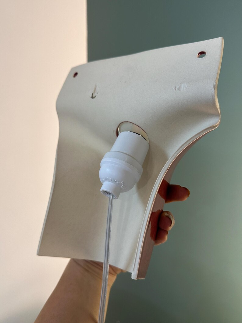 Pode incluir: Um candeeiro de parede em cer&acirc;mica branca com uma forma curva e alargada. A parte de tr&aacute;s apresenta um casquilho central de pl&aacute;stico branco com um cabo el&eacute;trico transparente que desce e dois pequenos orif&iacute;cios circulares de montagem.