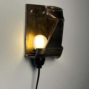 May include: A wall-mounted sconce featuring a dark, metallic, folded rectangular backplate with a smooth, reflective finish. A single, round, frosted white light bulb is positioned in the center, emitting a warm glow, connected to a black cord.