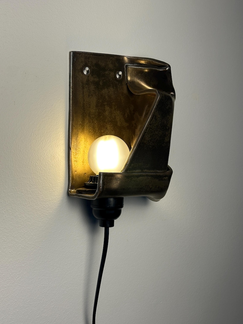 May include: A wall-mounted sconce featuring a dark, metallic, folded rectangular backplate with a smooth, reflective finish. A single, round, frosted white light bulb is positioned in the center, emitting a warm glow, connected to a black cord.