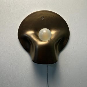 May include: A wall-mounted lamp featuring a circular, bronze-colored metallic shade with a soft, undulating, organic shape. A single, round, frosted white light bulb is positioned in the center of the shade, and a thin power cord hangs downward.