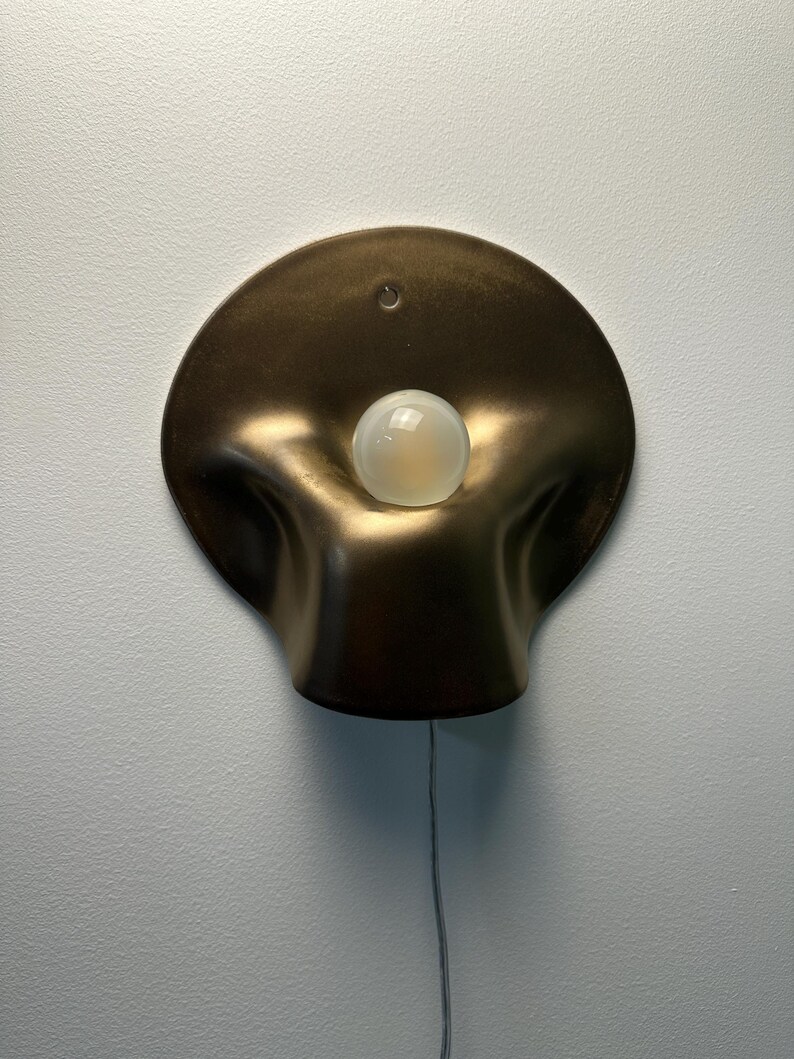 May include: A wall-mounted lamp featuring a circular, bronze-colored metallic shade with a soft, undulating, organic shape. A single, round, frosted white light bulb is positioned in the center of the shade, and a thin power cord hangs downward.