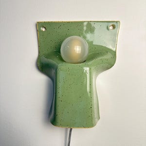 May include: A wall-mounted ceramic lamp with a glossy light green finish and dark speckles. The fixture features a wide, flat top section with two circular holes for mounting and a central, rounded protrusion holding a single spherical light bulb.