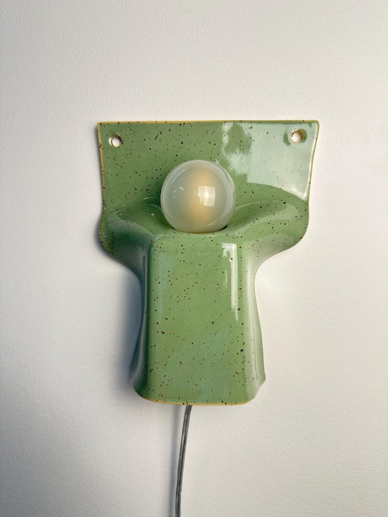 May include: A wall-mounted ceramic lamp with a glossy light green finish and dark speckles. The fixture features a wide, flat top section with two circular holes for mounting and a central, rounded protrusion holding a single spherical light bulb.