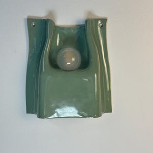 May include: A sage green ceramic wall light featuring a smooth, undulating, folded form with a central circular cutout. A single frosted white spherical light bulb is positioned within the recessed centre, and two metal grommets are visible at the top.
