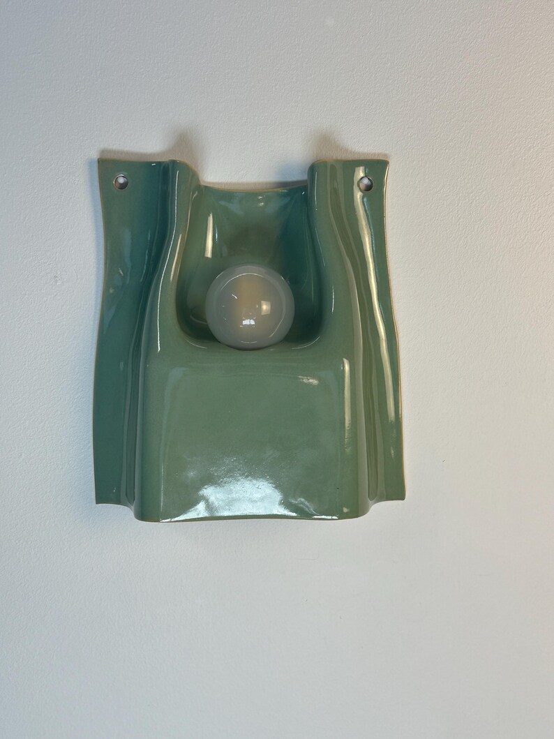 May include: A sage green ceramic wall light featuring a smooth, undulating, folded form with a central circular cutout. A single frosted white spherical light bulb is positioned within the recessed centre, and two metal grommets are visible at the top.