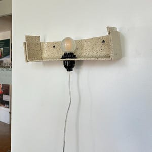 May include: A speckled beige wall sconce with a curved design. The sconce holds a light bulb with a black base and a clear cord. The sconce is mounted on a white wall.