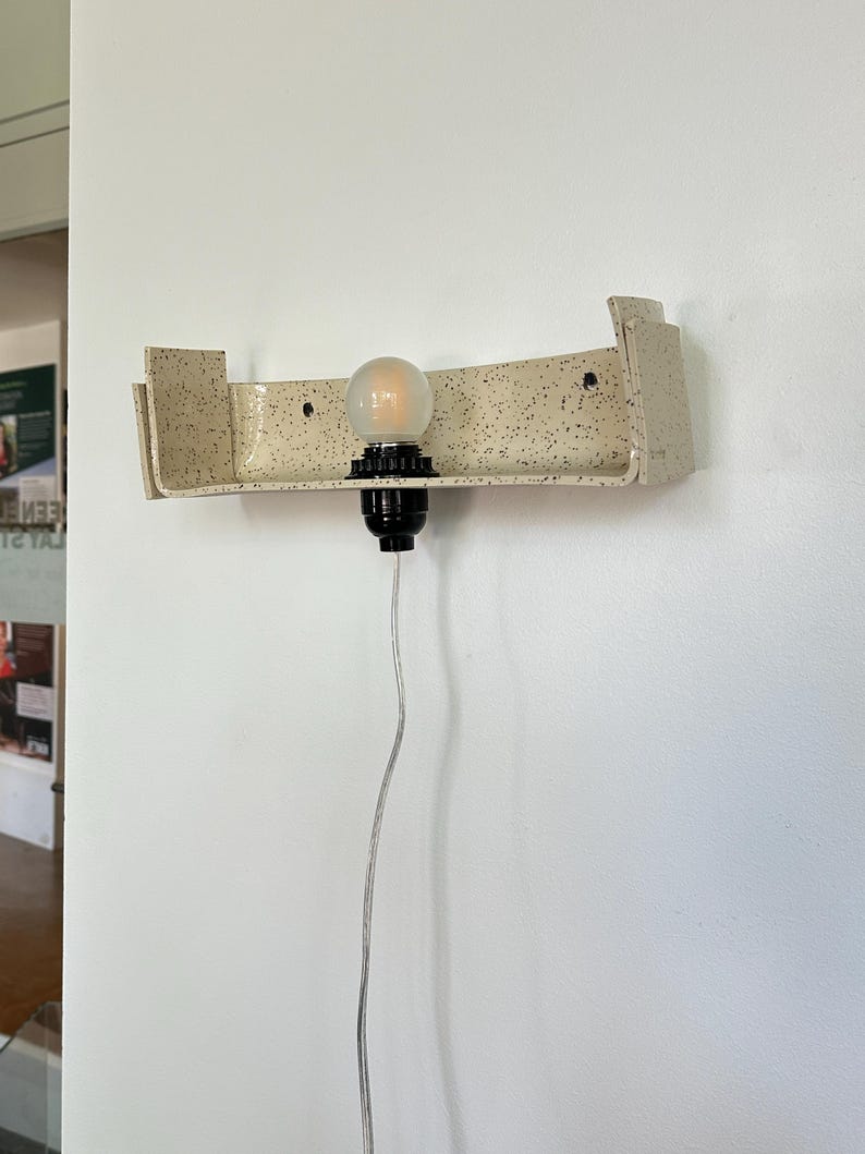 May include: A speckled beige wall sconce with a curved design. The sconce holds a light bulb with a black base and a clear cord. The sconce is mounted on a white wall.