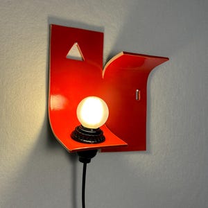 May include: A contemporary red wall light with an unusual, angular shape. It has a small, triangular cut-out and a curved shelf supporting a round light bulb. A black cable extends from the base, and the light is mounted on a grey wall.