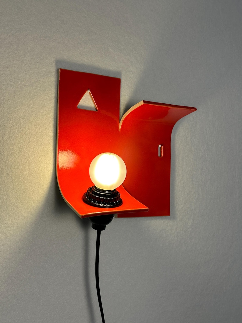 May include: A contemporary red wall light with an unusual, angular shape. It has a small, triangular cut-out and a curved shelf supporting a round light bulb. A black cable extends from the base, and the light is mounted on a grey wall.