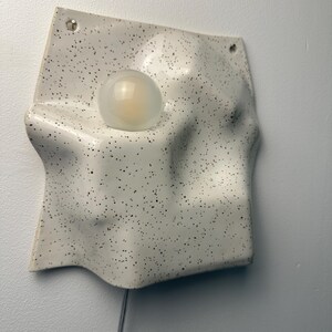 May include: A wall-mounted light fixture featuring a white ceramic base with a black speckled pattern. The base has an irregular, undulating surface with a circular cutout holding a single, round, frosted glass light bulb. A thin cord hangs below.