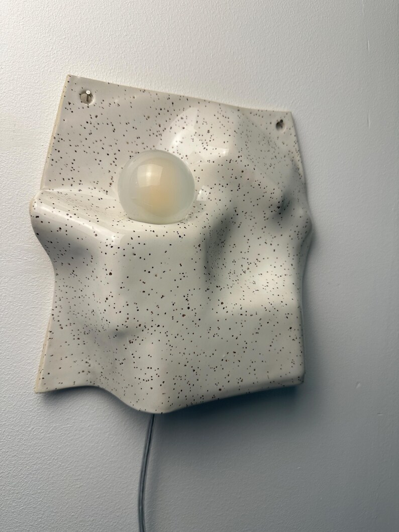 May include: A wall-mounted light fixture featuring a white ceramic base with a black speckled pattern. The base has an irregular, undulating surface with a circular cutout holding a single, round, frosted glass light bulb. A thin cord hangs below.