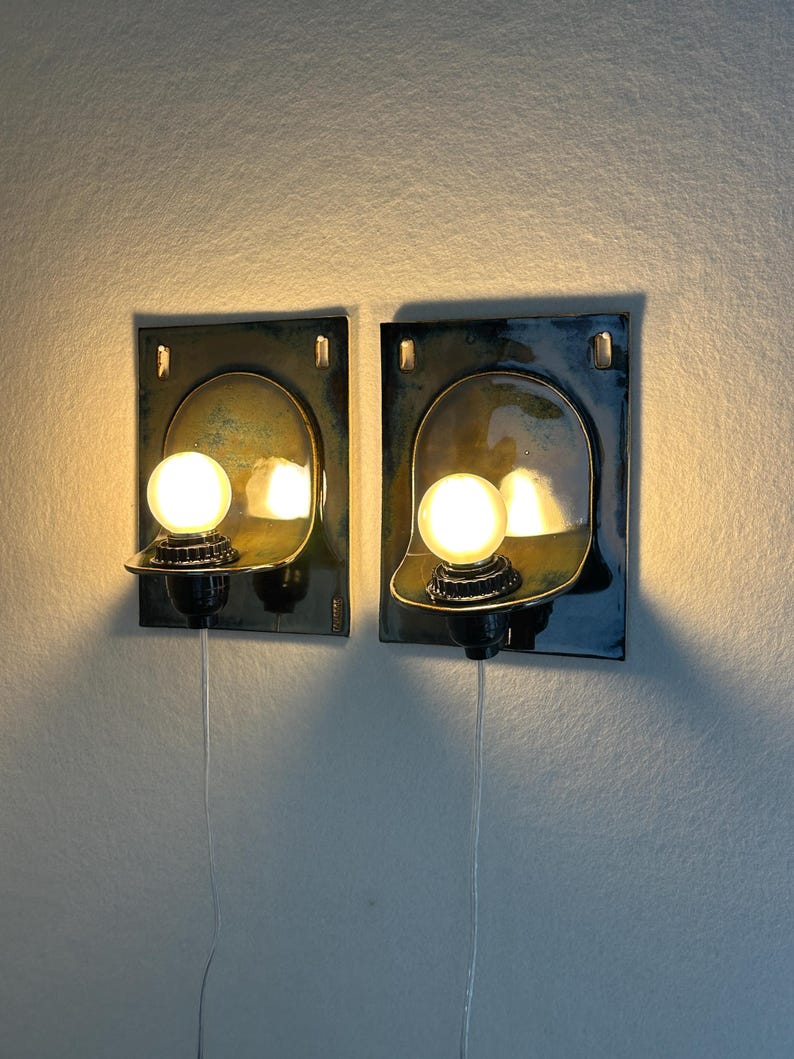 May include: Two wall-mounted sconces with a dark blue and gold finish. Each sconce features a rounded light fixture with a spherical bulb. The sconces are rectangular with a curved top and a small shelf to hold the bulb. The sconces are wired.