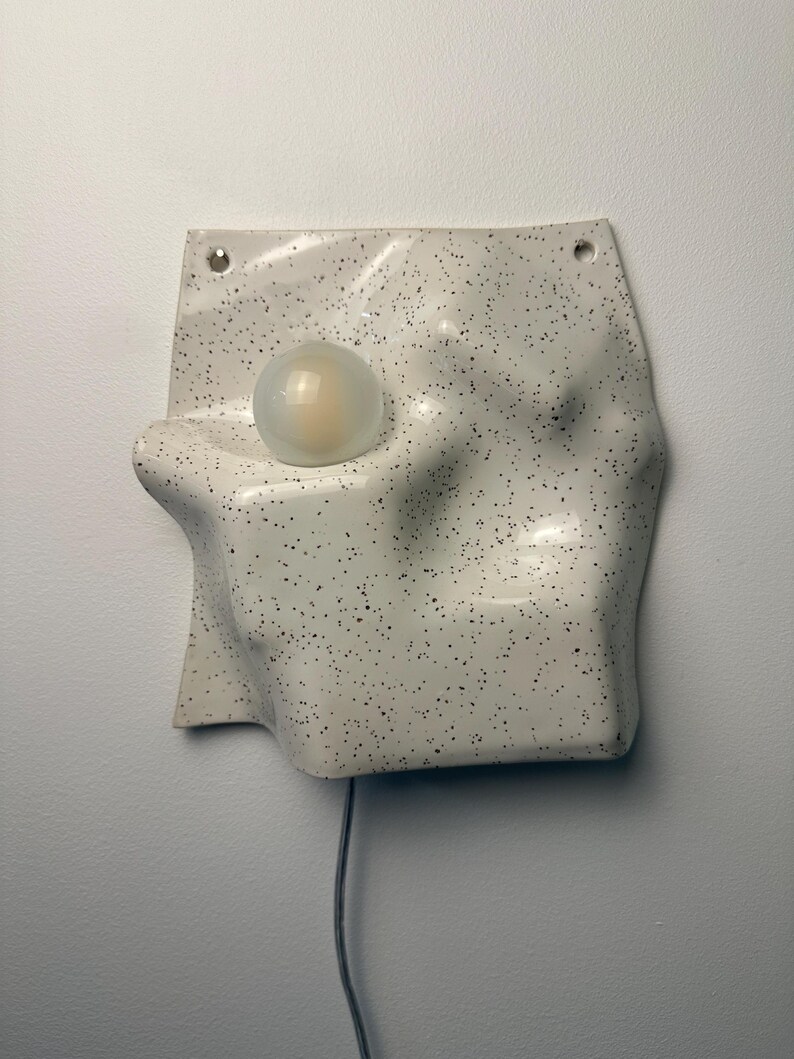 May include: A wall-mounted light fixture featuring a white ceramic base with a black speckled pattern. The base has an irregular, undulating surface with two circular holes at the top corners and a single frosted glass globe bulb attached.