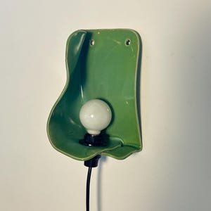Pu&ograve; includere: Una lampada da parete con un paralume in ceramica verde lucido dalla forma organica con bordi irregolari e ondulati. Una lampadina bianca rotonda &egrave; centrata all'interno della forma in ceramica, fissata a un portalampada nero e un cavo nero.