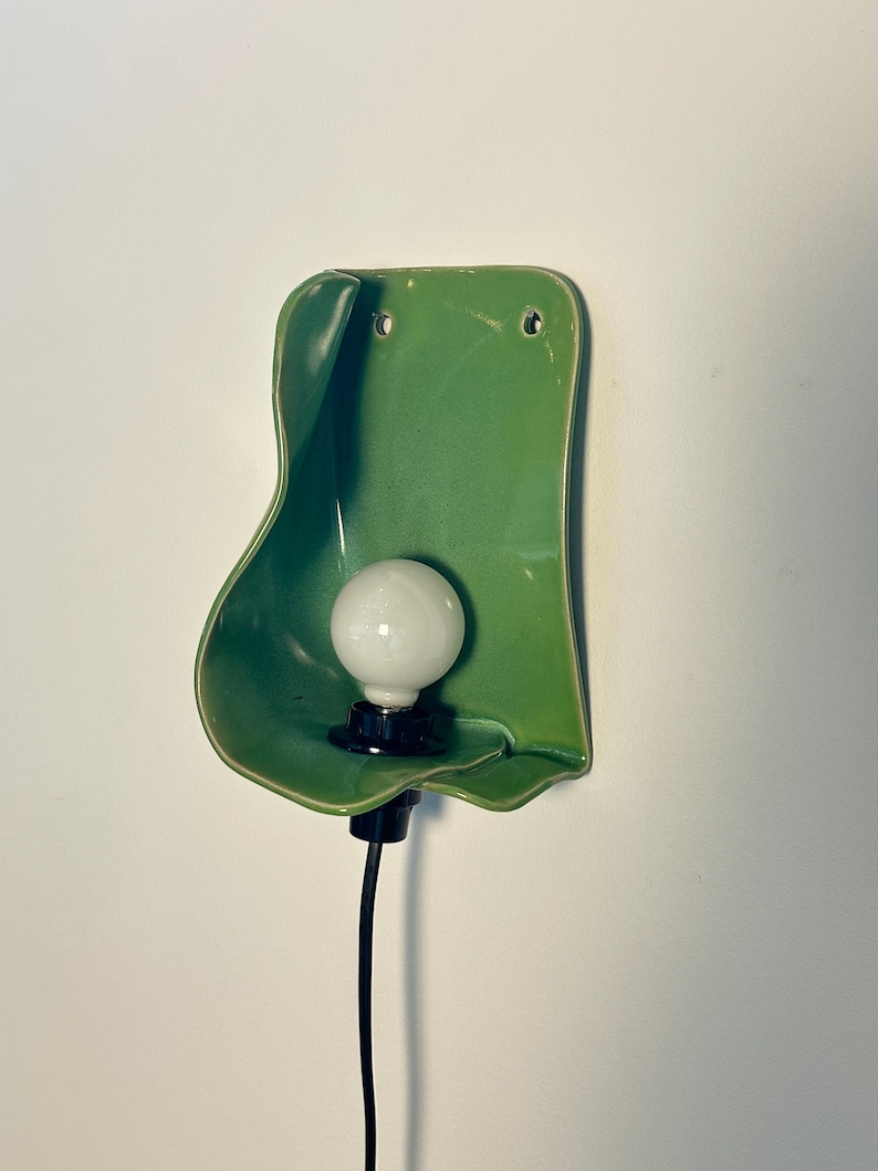 Pu&ograve; includere: Una lampada da parete con un paralume in ceramica verde lucido dalla forma organica con bordi irregolari e ondulati. Una lampadina bianca rotonda &egrave; centrata all'interno della forma in ceramica, fissata a un portalampada nero e un cavo nero.