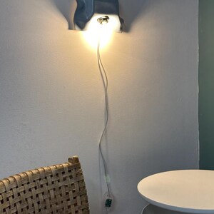May include: A wall-mounted light fixture with a wavy, light blue, molded shade that directs a warm, bright glow downward. The lamp features a thin white power cord that hangs vertically down the wall to a small, clear inline power switch.