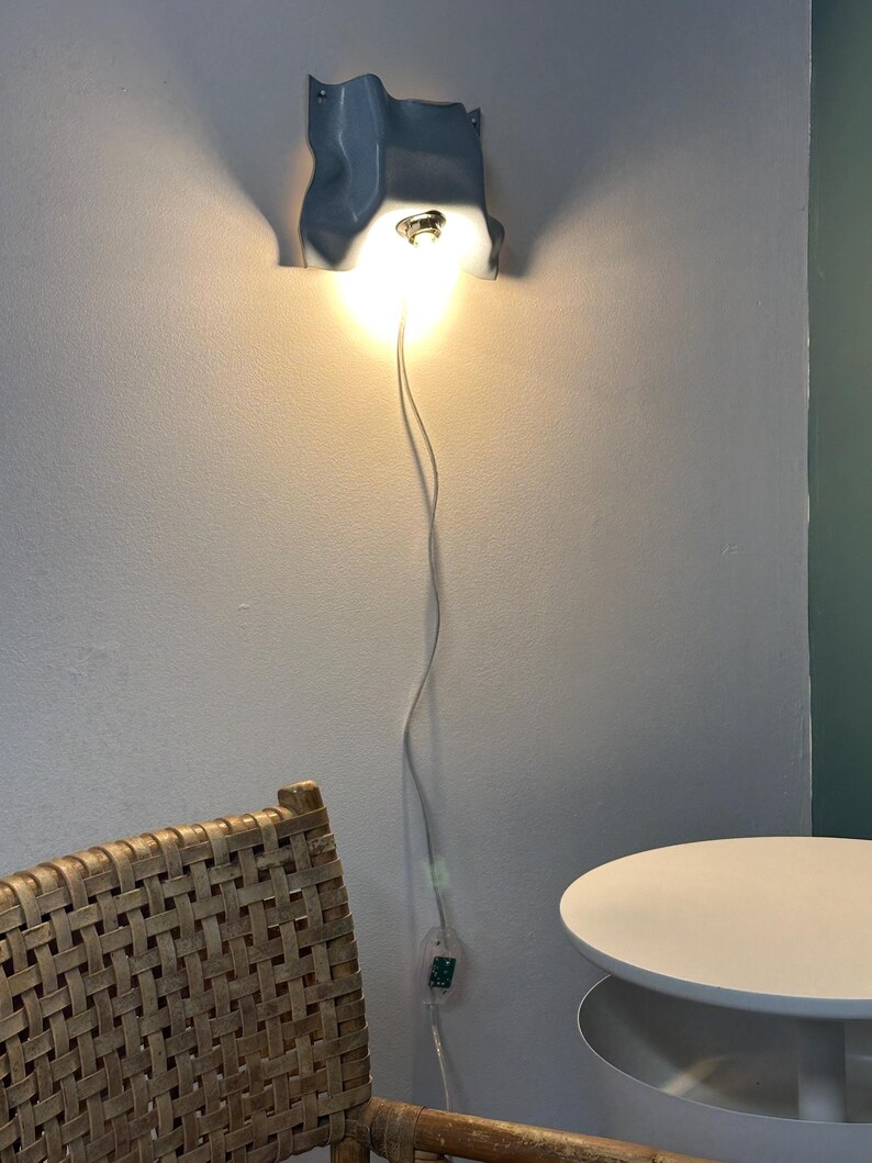 May include: A wall-mounted light fixture with a wavy, light blue, molded shade that directs a warm, bright glow downward. The lamp features a thin white power cord that hangs vertically down the wall to a small, clear inline power switch.