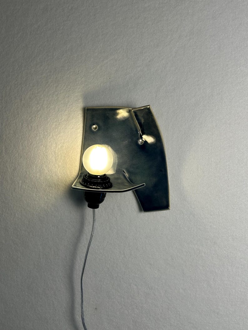 May include: A unique, abstract wall sconce with a dark metallic finish. The sconce features a small, round, illuminated light bulb and a black base. A silver cord extends downward from the base.