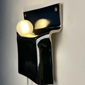 Peut inclure: Une applique murale compos&eacute;e d'une base en c&eacute;ramique noire brillante en forme de T, avec une ampoule sph&eacute;rique blanche lumineuse log&eacute;e dans la partie sup&eacute;rieure. Le luminaire pr&eacute;sente deux trous de fixation visibles aux coins sup&eacute;rieurs.