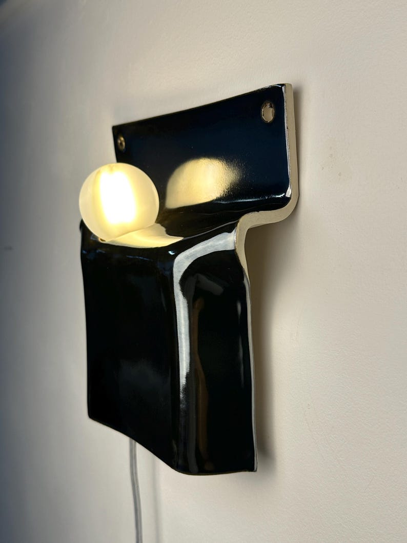 Peut inclure: Une applique murale compos&eacute;e d'une base en c&eacute;ramique noire brillante en forme de T, avec une ampoule sph&eacute;rique blanche lumineuse log&eacute;e dans la partie sup&eacute;rieure. Le luminaire pr&eacute;sente deux trous de fixation visibles aux coins sup&eacute;rieurs.