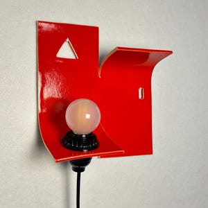 May include: A contemporary red wall light with a distinctive, geometric shape. It has a triangular aperture and a small shelf. A spherical, frosted light bulb is mounted on a black base, connected to a black cable.
