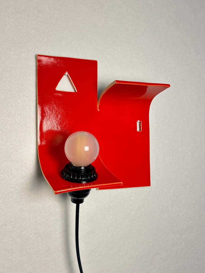 May include: A contemporary red wall light with a distinctive, geometric shape. It has a triangular aperture and a small shelf. A spherical, frosted light bulb is mounted on a black base, connected to a black cable.