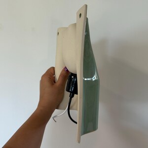 May include: A wall-mounted light fitting featuring a matte cream-coloured ceramic base with a smooth, glossy sage green front panel. The fitting includes a black socket and a black and white electrical cord extending from the back of the unit.