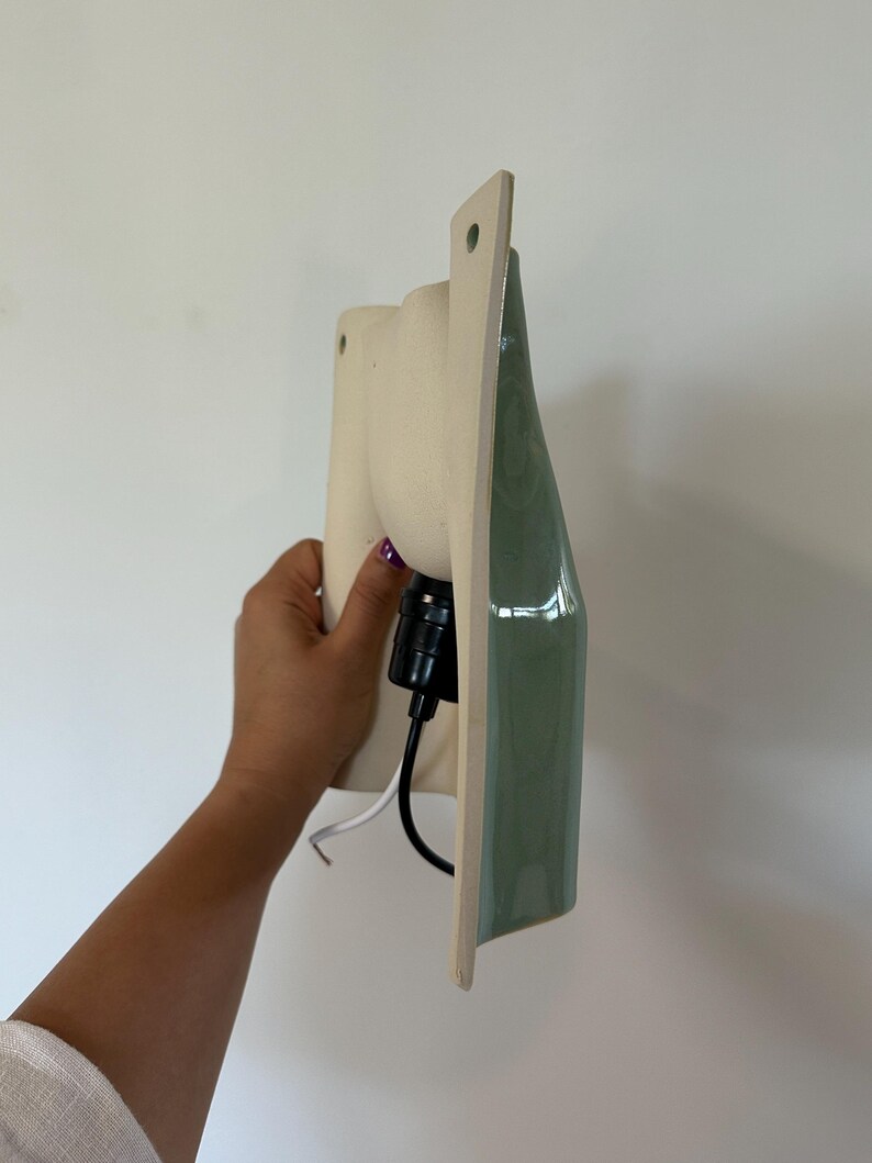 May include: A wall-mounted light fitting featuring a matte cream-coloured ceramic base with a smooth, glossy sage green front panel. The fitting includes a black socket and a black and white electrical cord extending from the back of the unit.