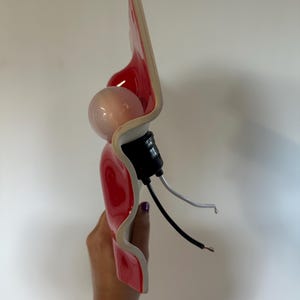 May include: A wall sconce featuring a glossy red, wavy ceramic shade with a white underside. The fixture includes a round light bulb, a black socket, and two exposed electrical wires, one black and one white, extending from the base.