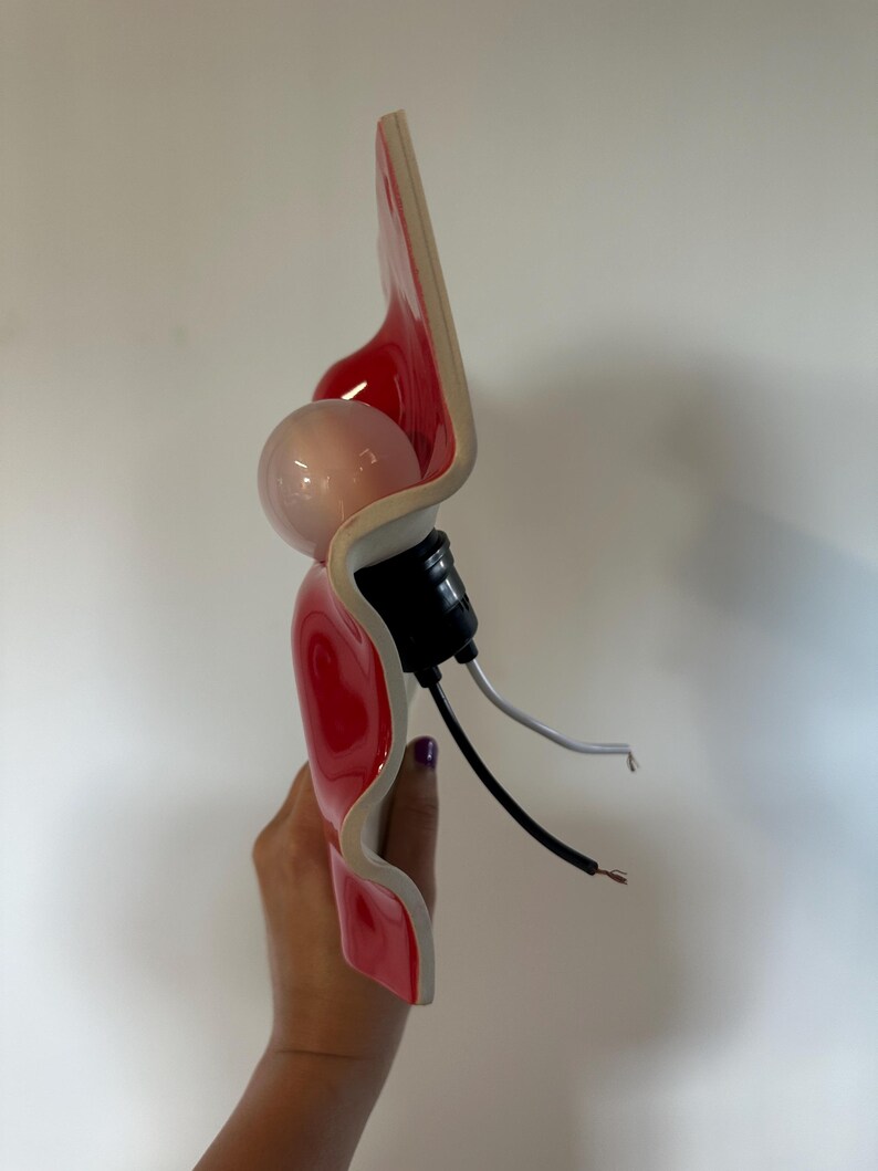 May include: A wall sconce featuring a glossy red, wavy ceramic shade with a white underside. The fixture includes a round light bulb, a black socket, and two exposed electrical wires, one black and one white, extending from the base.