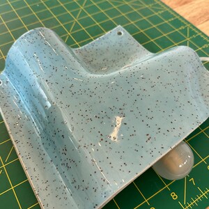 May include: A light blue ceramic wall sconce with a speckled dark brown finish. The fixture features a curved, angular design with two small circular mounting holes at the top corners and a single white light bulb socket visible underneath.