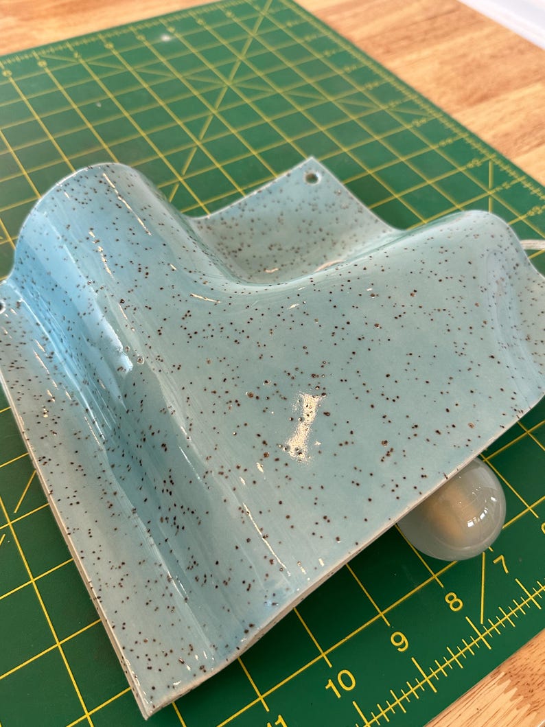 May include: A light blue ceramic wall sconce with a speckled dark brown finish. The fixture features a curved, angular design with two small circular mounting holes at the top corners and a single white light bulb socket visible underneath.