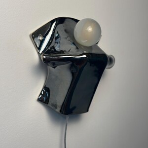 May include: A wall-mounted light fixture featuring a glossy, dark charcoal-colored ceramic base with an irregular, folded shape. A single, spherical, frosted white glass bulb is attached to the upper section, and a thin power cord hangs downward.
