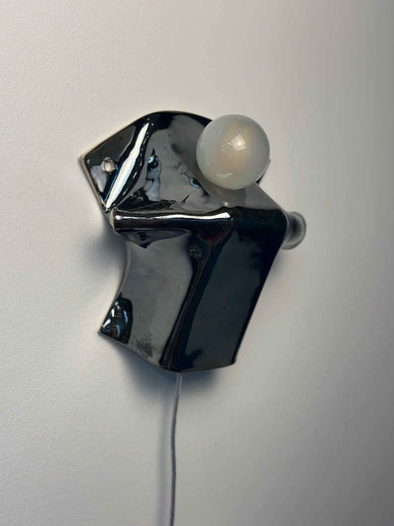 May include: A wall-mounted light fixture featuring a glossy, dark charcoal-colored ceramic base with an irregular, folded shape. A single, spherical, frosted white glass bulb is attached to the upper section, and a thin power cord hangs downward.