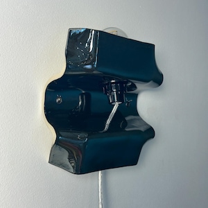 May include: A wall-mounted lamp featuring a glossy, dark teal ceramic fixture with an irregular, organic shape. The lamp includes a central black socket holding a clear light bulb, with a thin, transparent power cord extending downward from the base.
