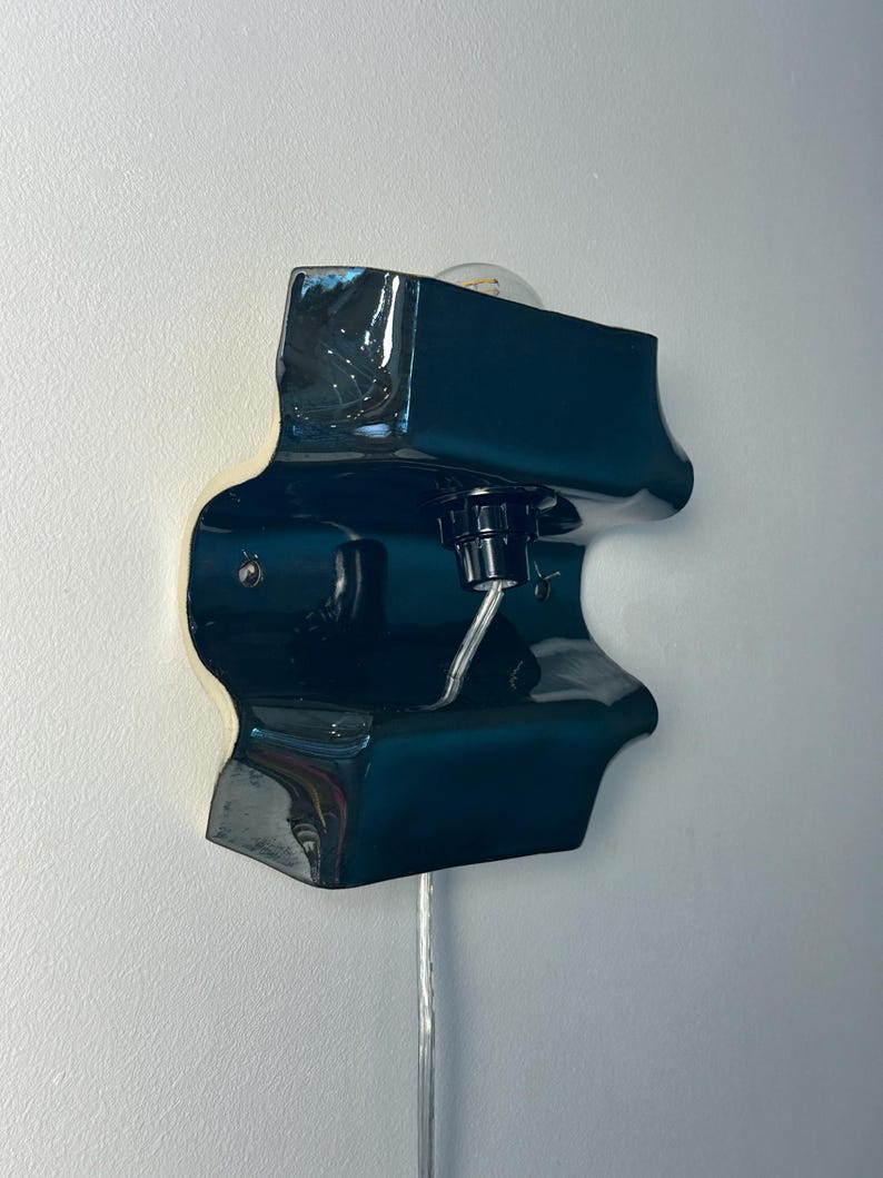 May include: A wall-mounted lamp featuring a glossy, dark teal ceramic fixture with an irregular, organic shape. The lamp includes a central black socket holding a clear light bulb, with a thin, transparent power cord extending downward from the base.